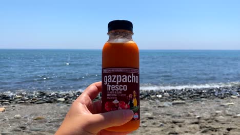 Halten-Sie-Eine-Frische-Traditionelle-Gazpacho-Miniflasche-Aus-Dem-Spanischen-Mercadona-Supermarkt-Am-Sonnigen-Strand-Von-Estepona,-Spanien,-Mit-Meerblick,-Kalte-Suppe-Für-Unterwegs,-4K-Aufnahme