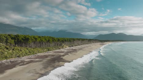 Panoramic-aerial-view-of-long,-wild-and-rugged-sandy-coastline-covered-in-driftwood-with-windswept-native-rimu-trees-of-Bruce-Bay-in-South-Westland,-New-Zealand-Aotearoa