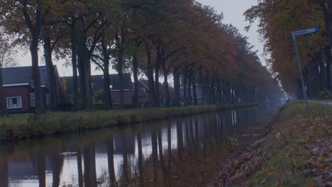 Water-canal-in-the-village-of-Netherlands-Holland-in-the-fall-autumn-with-houses,-trees-and-leaves-next-to-the-quiet-street-road