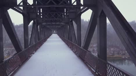 Cold-monochrome-aerial-along-snowy-abandoned-steel-bridge-deck,-winter