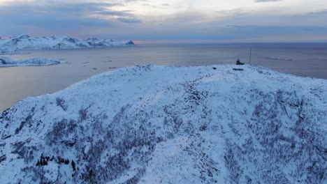 Drone-view-in-Tromso-area-in-winter-flying-over-a-snowy-mountain-peak-with-the-sea-on-the-horizon-in-Norway