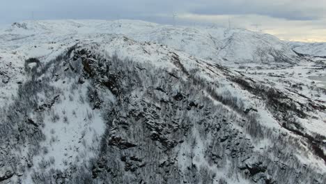 Drohnenansicht-In-Der-Gegend-Von-Tromsø-Im-Winter,-Die-über-Einen-Schneebedeckten-Berggipfel-Fliegt,-Umgeben-Vom-Meer-In-Norwegen-Und-Windmühlen-Am-Horizont