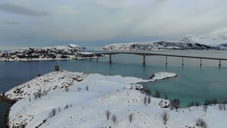 Drone-view-in-Tromso-area-in-winter-flying-over-a-small-island-an-under-a-bridge-connecting-two-islands-full-of-snow-over-the-sea-in-Norway