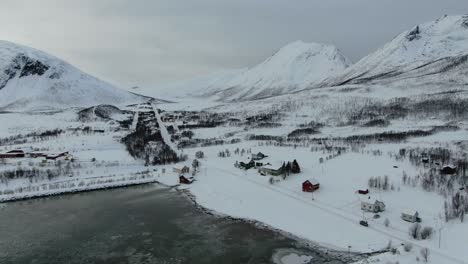 Drone-view-in-Tromso-area-in-winter-flying-over-a-snowy-landscape-surrounded-by-the-sea-and-a-frozen-port-with-boats-and-white-mountains-in-Norway