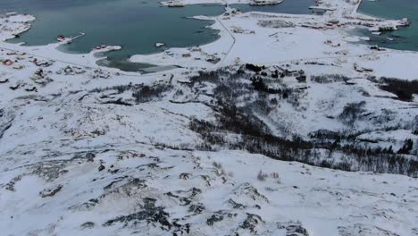 Drone-view-in-Tromso-area-in-winter-flying-over-a-snowy-mountain-peak-and-flat-islands-connected-by-bridges-with-small-towns-in-Norway