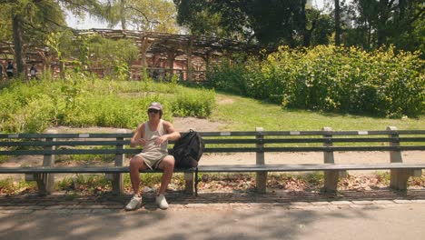 Man-sits-back-and-relaxes-at-bench-in-shade-at-central-park-new-york-city