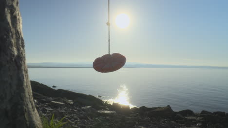 Rope-swing-moving-gently-in-the-breeze-on-the-shoreline-as-early-morning-autumn-sunlight-reflects-off-the-sea
