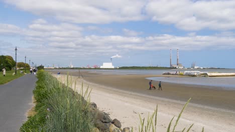 Gente-Caminando-En-Sandymount-Strand-Con-Vistas-A-La-Estación-Generadora-De-Poolbeg-Y-Al-Incinerador-De-Poolbeg-En-Dublín,-Irlanda