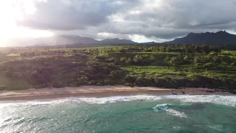 Aerial-view-over-clear,-transparent-ocean-water-foaming-at-the-shore,-washing-the-golden-sand-of-the-beach,-green-mountains-and-amazing-sunset-with-gorgeous-tropical-nature-of-Hawaii