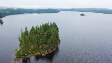 Epic-nature-scene-of-lush-green-pine-island-surrounded-by-calm-Norwegian-lake,-aerial
