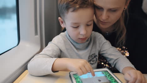 Child-playing-game-on-pad-when-traveling-by-train-with-mom