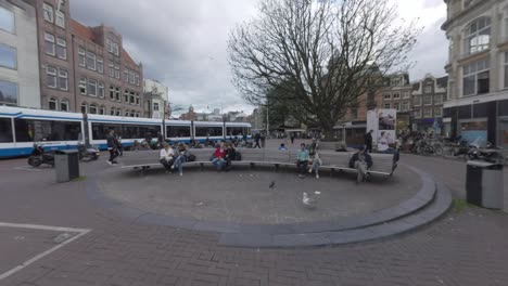 Koningsplein-with-people-on-benches-and-tram-passing-by-Amsterdam-Netherlands