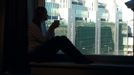 Woman-drinking-coffee-on-the-windowsill-with-city-view-in-window
