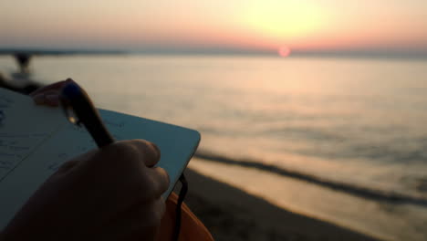 Frau-Macht-Sich-Notizen,-Während-Sie-Bei-Sonnenuntergang-Am-Meer-Sitzt