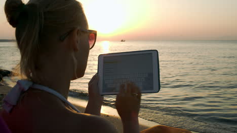 Frau,-Die-Bei-Sonnenuntergang-Am-Meer-Sitzt-Und-Auf-Einem-Pad-Tippt