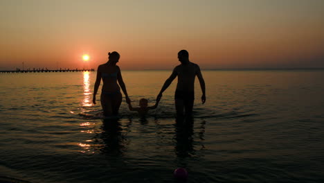 Family-silhouettes-coming-out-of-sea-at-sunset-three-people