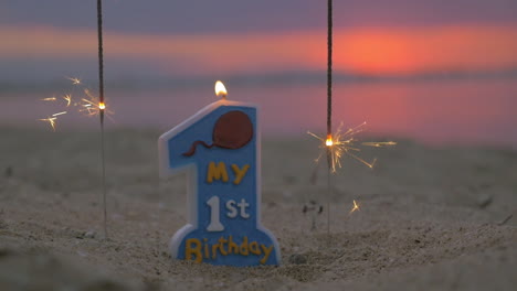 Slow-motion-view-of-candle-and-two-sparklers-standing-in-the-sand-on-beach-against-blurred-sunset