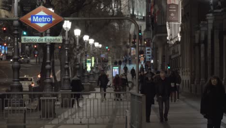 Vista-Nocturna-De-Gente-Caminando-Por-La-Acera-Con-Entrada-De-Metro-En-La-Noche-De-Madrid