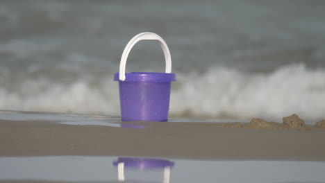 Ein-Lila-Spielzeugeimer-Im-Sand-Am-Strand-Mit-Glitzernden-Wellen-Im-Hintergrund