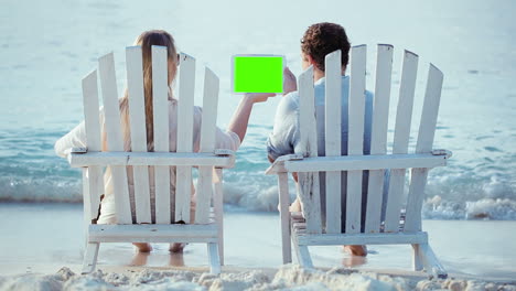 Woman-and-man-sitting-on-the-beach-looking-at-pad-with-green-screen