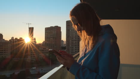 Frau-Mit-Smartphone-Auf-Dem-Balkon-In-Der-Stadt-Bei-Sonnenaufgang