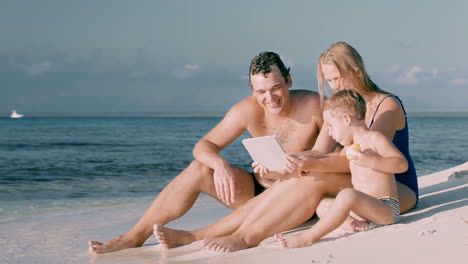 Happy-family-of-three-sitting-by-the-sea-and-using-pad