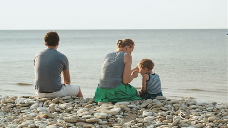 Family-of-three-sitting-on-pebble-beach-by-the-water