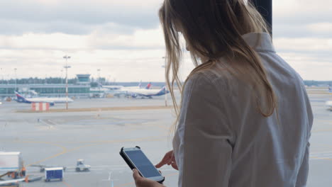 Frau-Schreibt-SMS-Auf-Dem-Handy-Und-Blickt-Auf-Den-Flughafenbereich