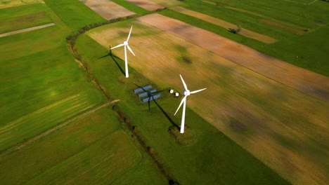 Drone-shot-of-two-working-wind-turbines-and-a-few-solar-panels-producing-green-electric-energy-on-a-cultivated-field-on-a-sunny-summer-day,-use-of-renewable-resources-of-energy,-still-shot