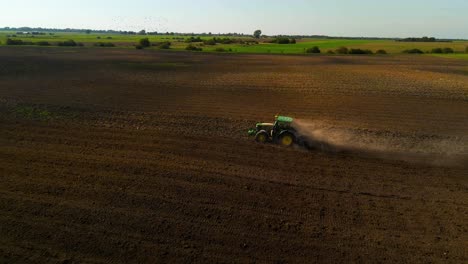 Drone-shot-of-a-green-John-Deere-tractor-preparing-the-field-for-plantation-with-a-cloud-of-dust-behind-tractor-on-a-sunny-summer-day,-parallax-shot