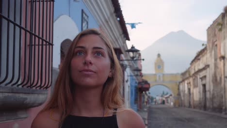 Young-beautiful-blonde-girl-with-blue-eyes-smiles-at-camera,-traveling-in-colonial-city-of-Antigua-Guatemala-with-cobblestone-streets-and-yellow-Santa-Catalina-clock-tower-visible-in-the-back