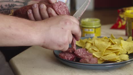 Close-up-shot-of-knife-cutting-salami-into-slices-and-dropping-onto-plate-with-corn-chips