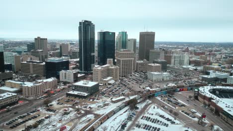 Heranzoomen-Beim-Aufbau-Einer-Städtischen-Winnipeg-Manitoba-Kanada-Innenstadt,-Wolkenkratzergebäude-In-Der-Stadt,-Bewölkter-Blauer-Himmel,-Landschaft,-Skyline,-Schneiender-Winter,-Drohne-4K-Aufnahme-Mit-Durchfahrendem-Bunten-Zug