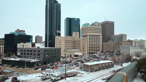 Establishing-Reveal-Shot-of-Winnipeg-Manitoba-Canada-Downtown-Skyscraper-Buildings-in-Overcast-Landscape-Skyline-Snowing-Winter-Drone-4k-Shot-with-Multicolored-Train-Passing-Through-City-Center