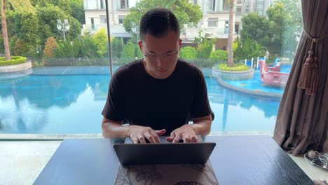 Asian-Millennial-Man-Nodding-Head-to-Music-While-Working-on-Laptop-in-his-Luxurious-Home-Overlooking-Pool