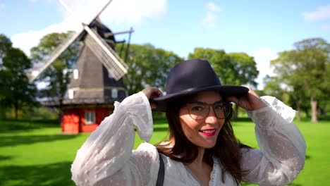 Mujer-Sonriendo-A-La-Cámara-Mientras-Se-Pone-Un-Sombrero-Y-Luego-Mira-El-Molino-De-Viento-En-Kastellet,-Dinamarca