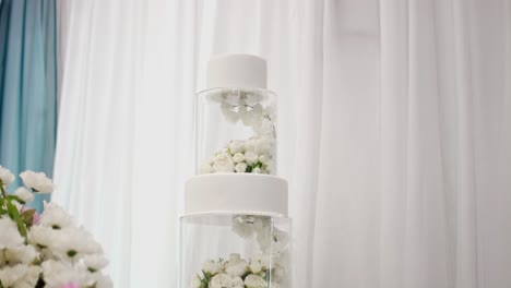 A-close-up-shot-of-a-3-tier-white-wedding-cake-with-acrylic-clear-cake-stands-with-flowers-inside-them-in-between-the-two-middle-layers