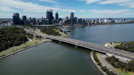 View-by-drone-tracking-towards-Perth,-Western-Australia,-flying-over-Narrows-Bridge-on-the-Swan-River,-train-crossing-the-bridge-between-both-lanes-of-traffic