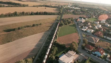 Aerial-drone-footage-of-a-train-emerging-from-a-vast-cornfield-and-entering-a-charming-town,-lush-greenery