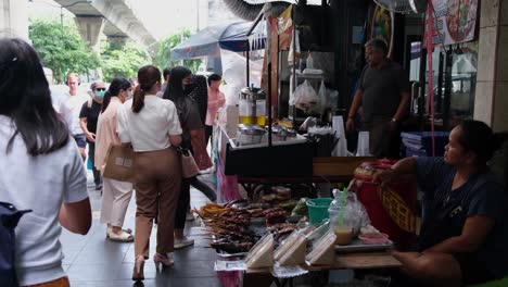 Eine-Verkäuferin,-Die-Sich-Von-Der-Hitze-Der-Morgensonne-Auf-Der-Straße-Luft-Zufächelt,-Während-Sie-Snacks-Und-Gegrilltes-Essen-An-Passanten-Verkauft,-Straßenessen-Entlang-Der-Sukhumvit-Road-In-Bangkok,-Thailand