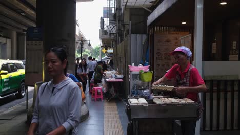 Woman-with-a-red-shirt-selling-broiled-bananas-skewed-together-on-a-sidewalk-and-people-come-and-go,-a-pharmacy-is-also-seen-at-the-background,-Street-Food-along-Sukhumvit-Road-in-Bangkok,-Thailand