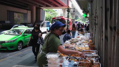 Verkäufer-Rührt-Und-Arrangiert-Lebensmittel-In-Behältern-Und-Bereitet-Sie-Für-Die-Mittagszeit-Entlang-Der-Sukhumvit-Straße-Vor,-Während-Der-Verkehr-Voranschreitet,-Straßenessen-Entlang-Der-Sukhumvit-Straße-In-Bangkok,-Thailand