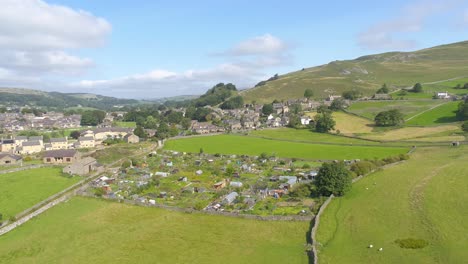 Drone-footage-moving-sideways,-panning-and-ascending-revealing-the-hills-and-countryside-around-the-village-of-Settle,-Yorkshire,-UK,-including-farmland,-houses,-fields,-stone-walls-and-allotments