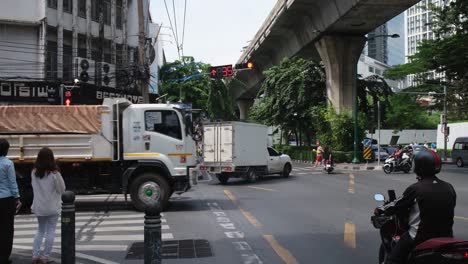 Grab-motorcycle-delivery-passes-by,-motor-taxi-with-a-woman-using-her-mobile-phone-while-others-wait-fro-the-green-walk-light-sign-for-everybody-to-cross-a-busy-street-in-Sukhumvit,-Bangkok,-Thailand