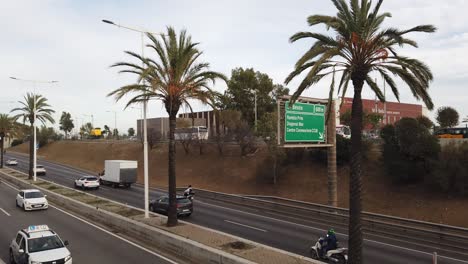 Panorámica-De-La-Autopista-Mediterránea,-Coches-Circulando-Por-La-Playa-De-Barcelona,-España,-Con-Palmeras-Ondeando-Con-El-Viento.