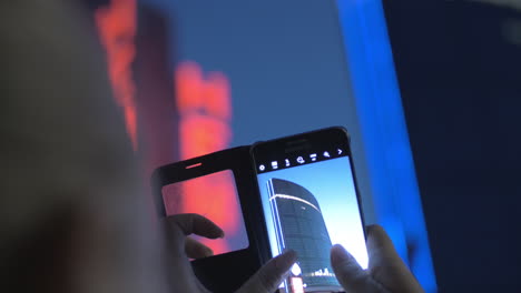 Close-up-view-of-woman-taking-a-picture-of-huge-blue-skyscraper-by-smartphone-against-evening-sky-Rotterdam-Netherlands