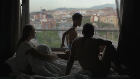 Vista-De-La-Familia-En-La-Cama-Con-Un-Hijo-Pequeño-Contra-Una-Enorme-Ventana-Panorámica-Y-El-Paisaje-Urbano-De-Barcelona,-España