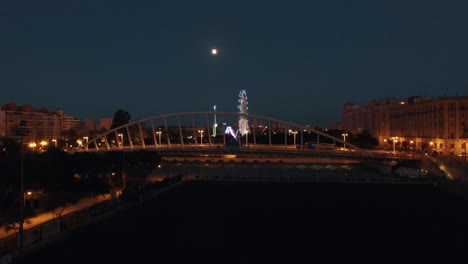 Aerial-night-view-of-lighted-ferris-wheel-and-bridge-against-sky-with-moon--Valencia-Spain