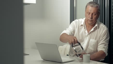 Un-Hombre-Adulto-Serio-Trabajando-En-Una-Computadora-Portátil-Mientras-Toma-Café-En-La-Cocina