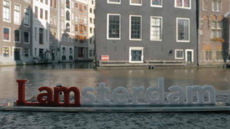 Blick-Auf-Die-Kleine-Plastikfigur-Der-Iamsterdam-Buchstabenskulptur-Auf-Der-Brücke-Vor-Verschwommenem-Stadtbild-In-Amsterdam,-Niederlande